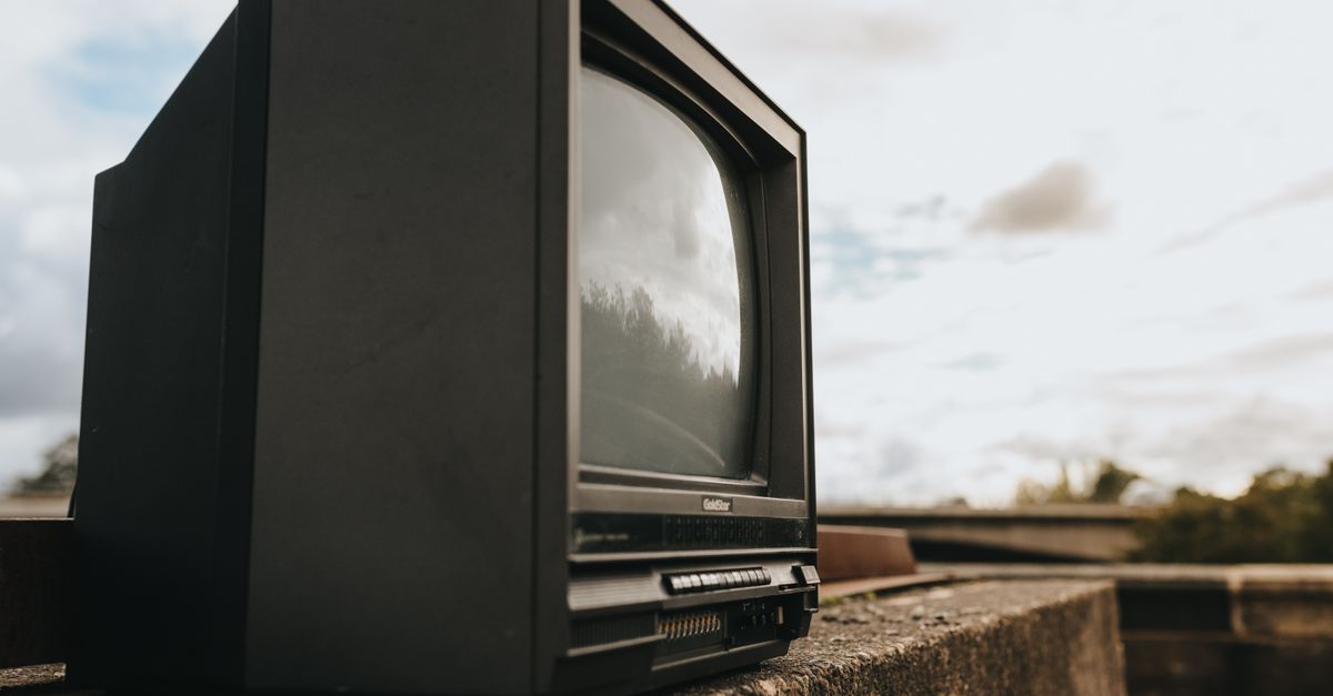 Petite télévision noire vintage posée sur muret de pierre, ciel nuageux et rue floue en arrière-plan