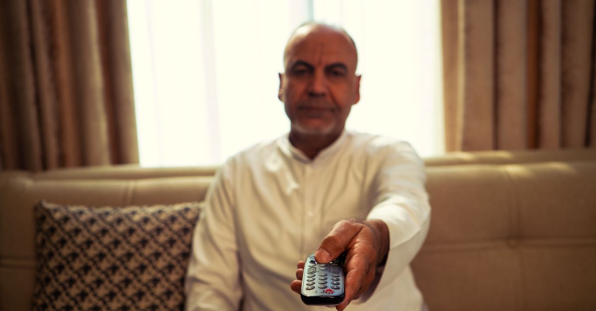 Homme âgé assis dans son salon, tendant une télécommande vers la télévision allumée