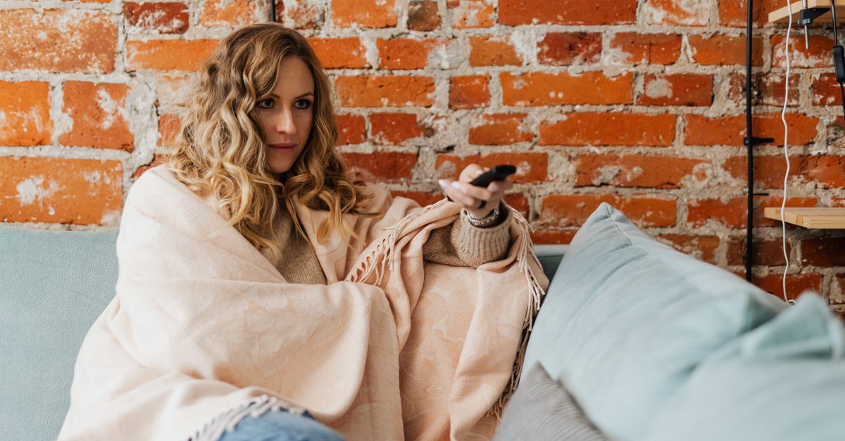 Femme emmitouflée dans une couverture tenant une télécommande devant la télévision allumée