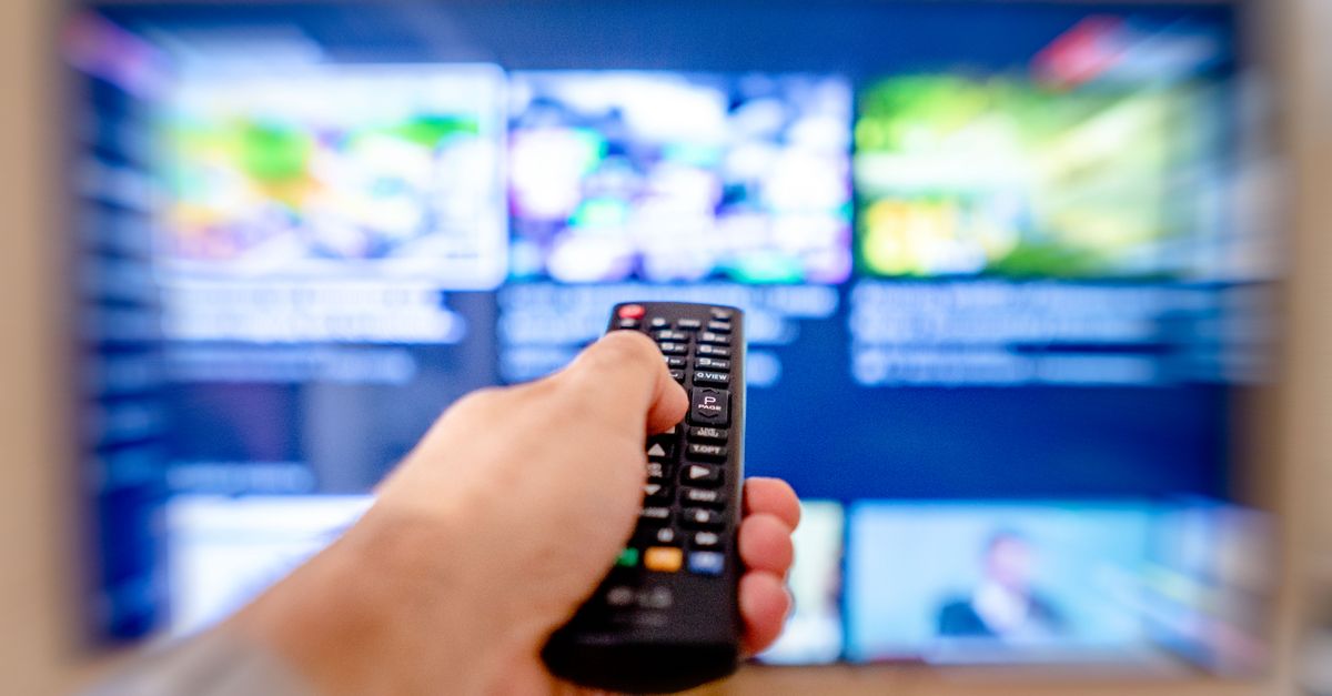 Main tenant une télécommande dirigée vers un écran de télévision flou dans un intérieur chaleureux