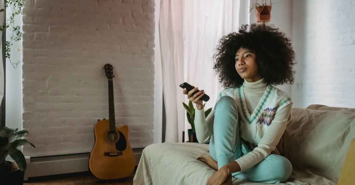Jeune femme assise sur un canapé, zappant des chaînes avec une télécommande à la main.