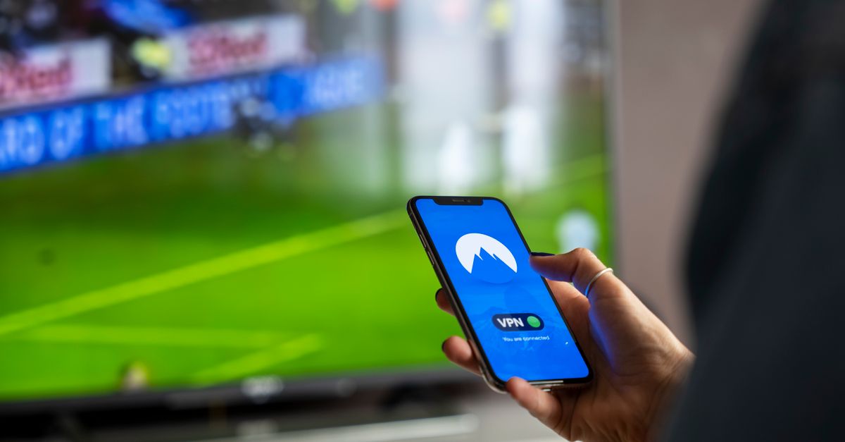 Zoom sur une main tenant un smartphone affichant une application VPN, match sportif diffusé à la télévision