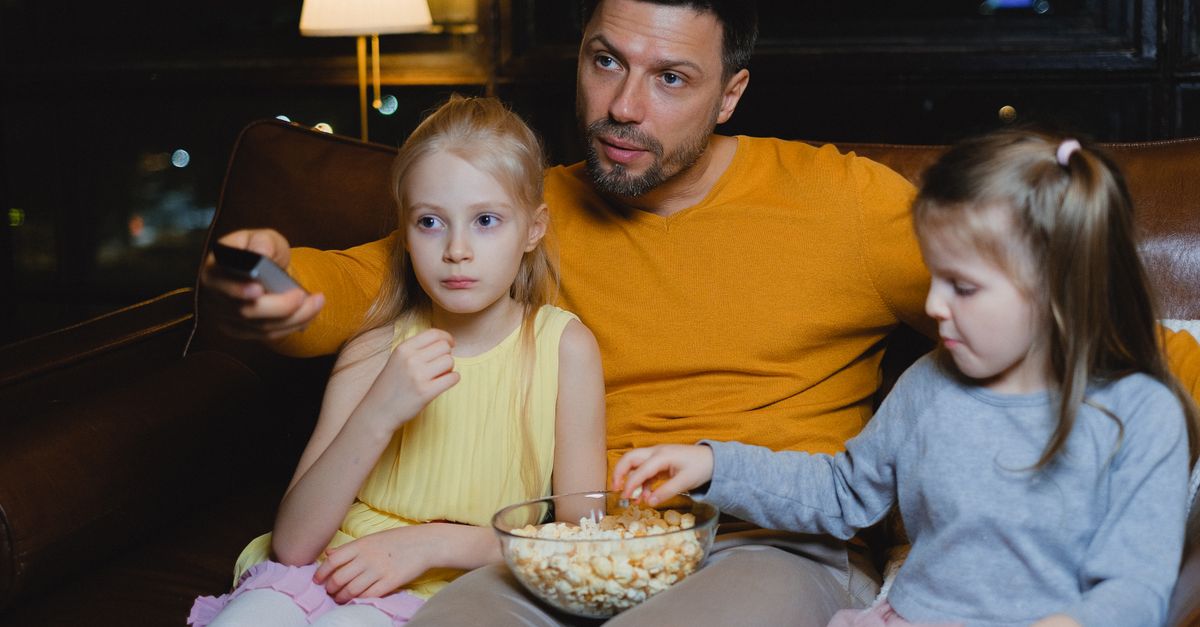 Père et ses deux filles assis sur le canapé partageant du popcorn pendant une soirée cinéma à la maison.