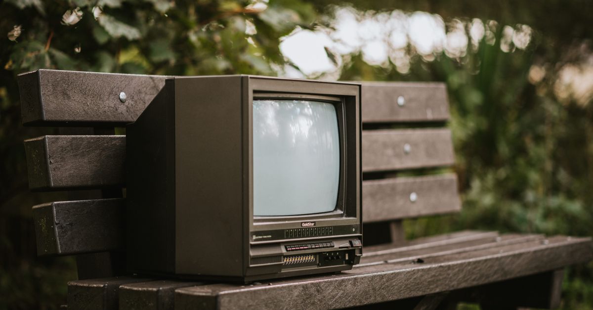 Petite télévision ancienne noire posée sur un banc près d’un buisson verdoyant en campagne.