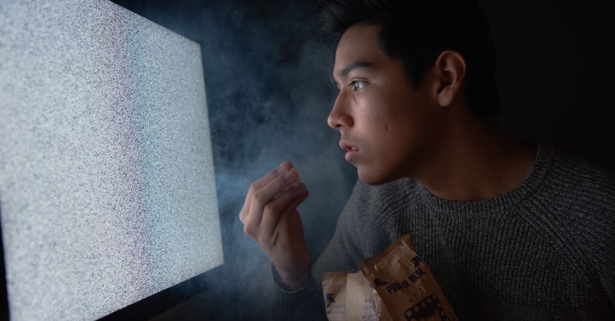 Jeune homme assis dans une pièce sombre, observant des parasites à l’écran en mangeant du popcorn.
