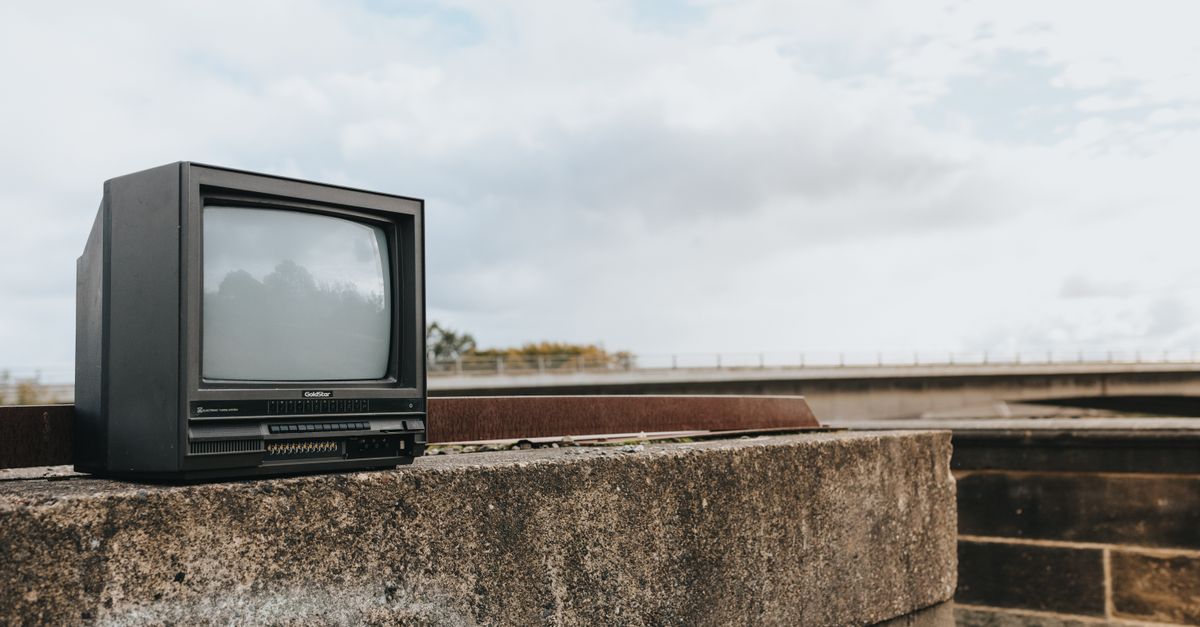 Télévision rétro noire installée sur une bordure en béton taguée, pont visible au loin sous un ciel nuageux.