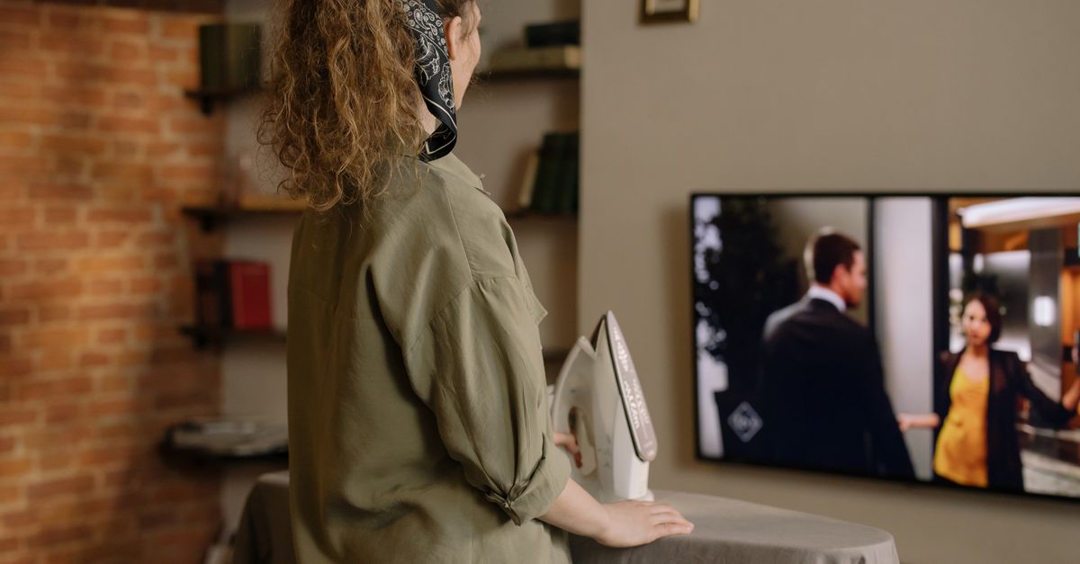 Femme repassant du linge tout en regardant la télévision dans un salon chaleureux aux murs en briques.