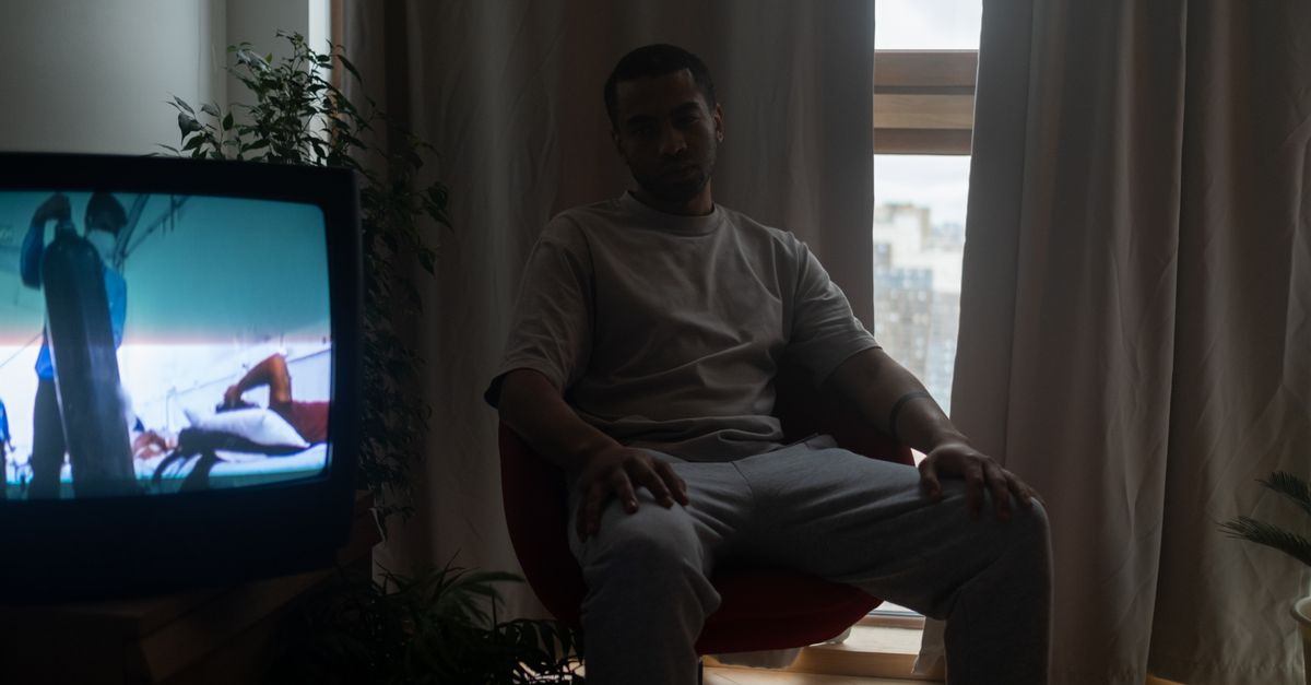 Homme assis dans une pièce sombre regardant la télévision, rideaux tirés derrière lui