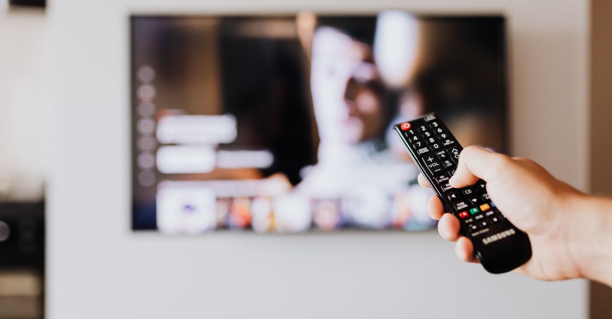 Main tenant une télécommande devant un écran de télévision flou en arrière-plan.