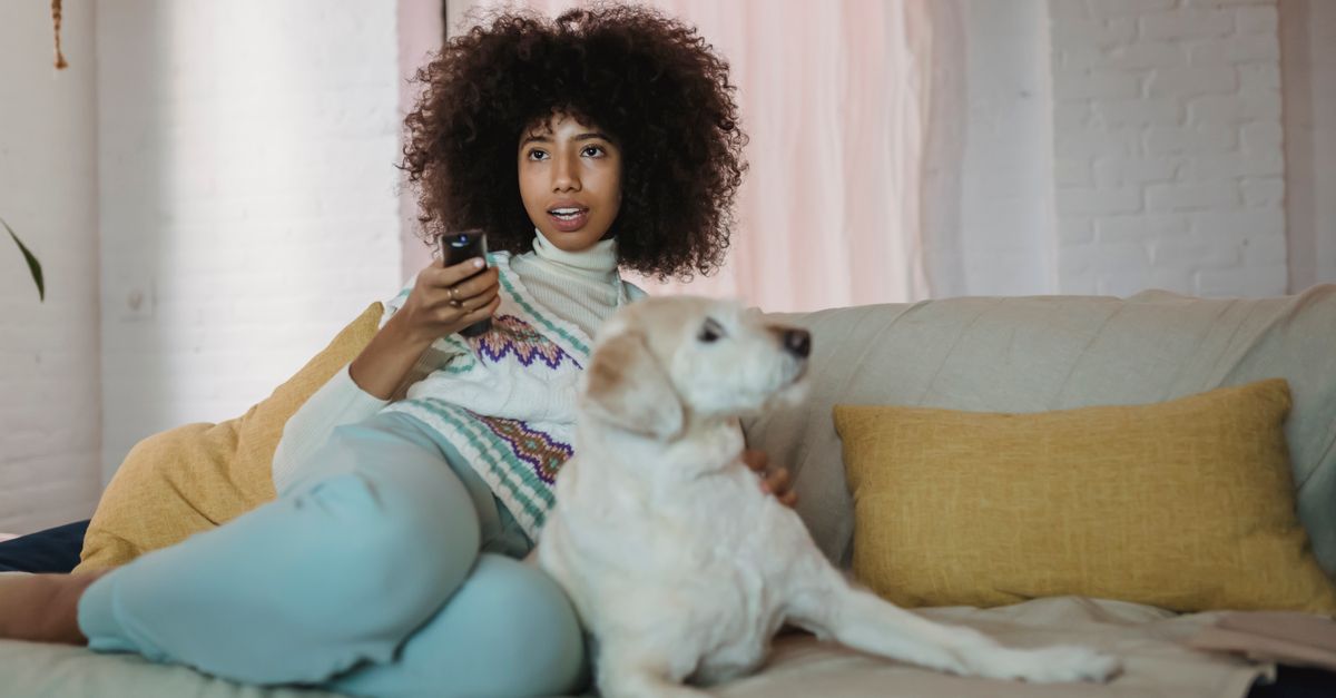 Jeune femme afro-américaine câlinant son chien sur un canapé en regardant la télévision.