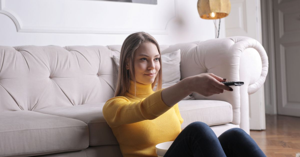 Femme détendue en pull jaune assise sur un canapé, télécommande à la main devant la télévision