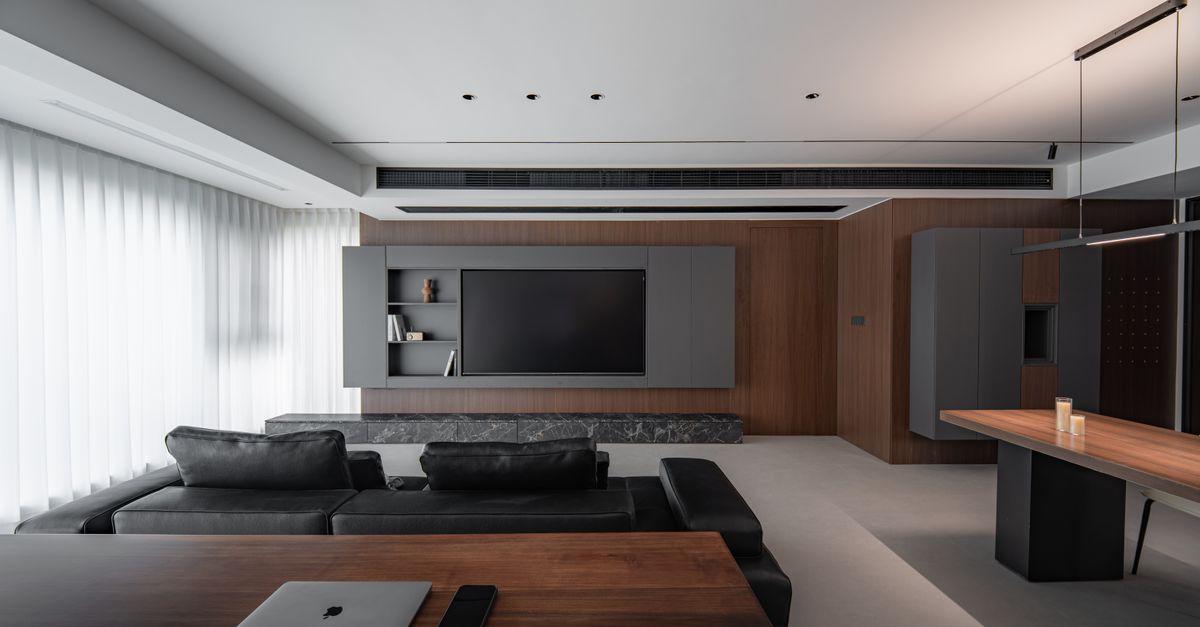 Grand salon moderne avec canapés noirs et bureau en bois près d’une télévision allumée